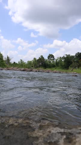 Serene River Flow with Lush Greenery and Cloudy Sky 

A tranquil river flows over rocks under a bright, cloudy sky, surrounded by dense tropical forest. A peaceful scene of nature