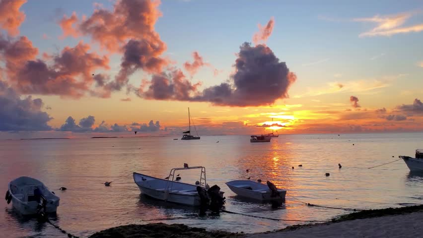 Scenic view tropical sunset over calm ocean waters, with small motorboats anchored near shoreline and sailboats visible on the horizon. Warm golden light reflects on sea surface