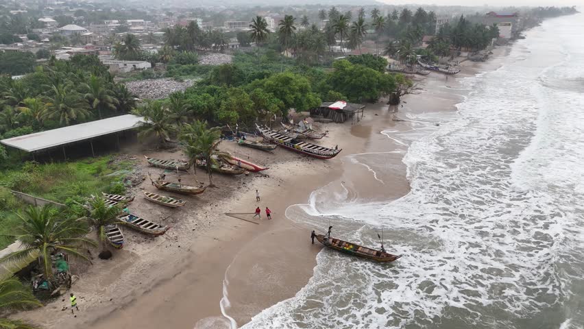 High-angle drone shot of the Atlantic shoreline at Accra Beach, Ghana. White waves crashing on the sandy beach with palm trees and coastal buildings in the background