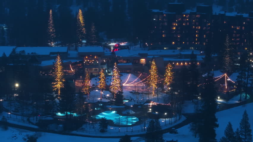 Drone top view of mountain ski resort hotel at night. Snow covered buildings, glowing festive trees and outdoor heated pool create warm winter travel atmosphere.