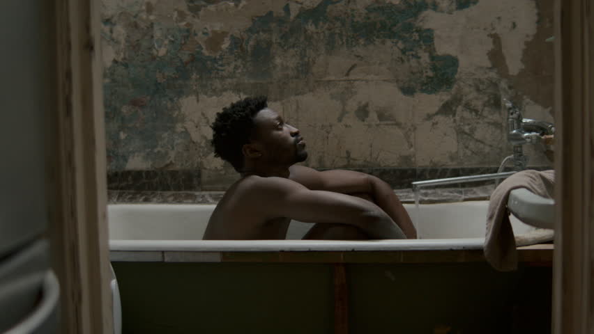 Young Black man sitting inside bathtub, resting his head on arms, feeling exhaustion or suffering from depression in worn bathroom with textured wall. Side view, zoom in shot