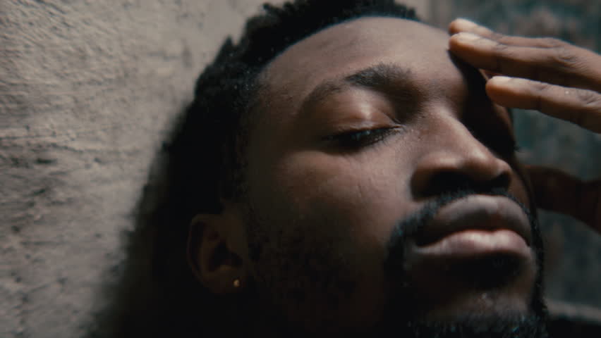 Close up view of young Black man rubbing his face with hands while leaning against rough textured wall in bathroom, struggling with depression or emotional burnout