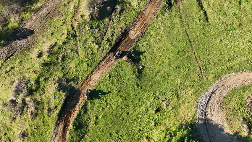 Cinematic top-down aerial shot tracking two ADV motorcycles as they ride along a winding dirt path through a vibrant green field in Chile.