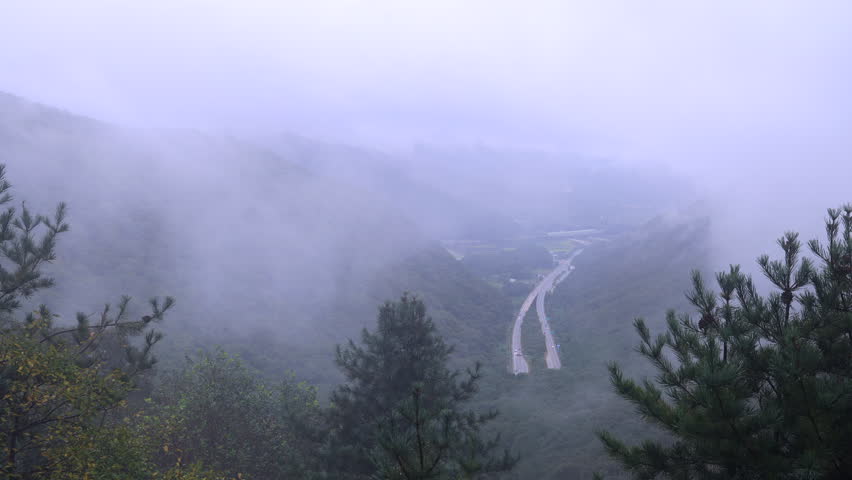A divided highway curves through a steep valley near Ihwaryeong Pass, with fog spilling between forested slopes and softening the distant ridges.