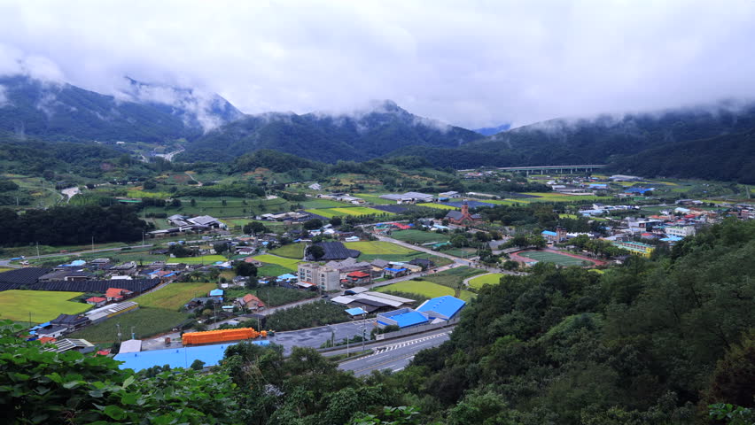 From Ihwaryeong Pass near Yeonpung Village, low clouds cling to ridgelines while rice fields, roofs, and village streets spread across the valley.