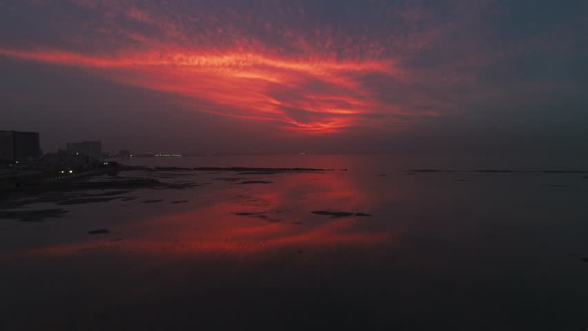 Wide view of a dramatic red sunset sky reflecting over calm coastal waters, with distant city skyline silhouettes and shoreline visible under evening light.