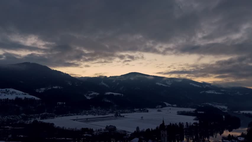 Cinematic timelapse of dramatic clouds moving over a snowy valley and mountain landscape during a glowing winter sunset.
