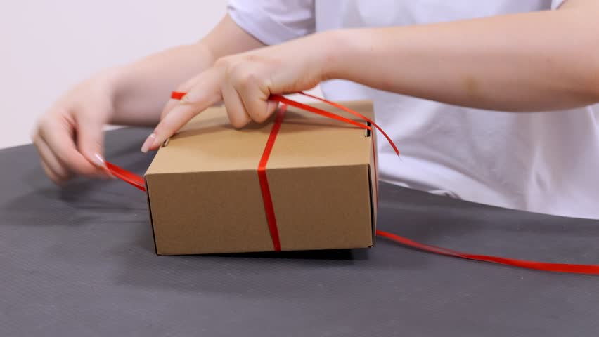 Person is tying a red ribbon around a brown box. The box is on a table. The person is wearing a white shirt