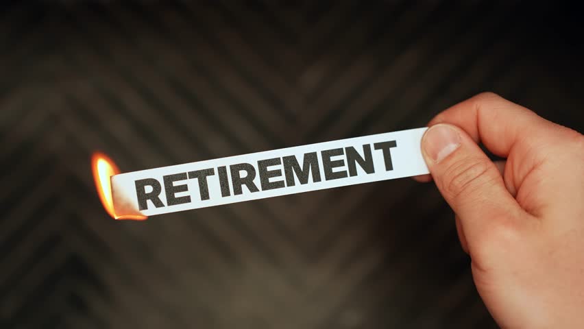 Close-up hand holds a paper strip reading “RETIREMENT” as the edge burns with a small flame, symbolizing financial risk, inflation, and uncertain future planning with copy space.