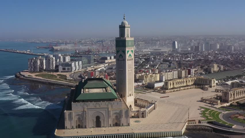 Close-up aerial drone shot highlighting the architectural details of the Hassan II Mosque minaret. Captures the iconic landmark against the backdrop of Casablanca's port and Atlantic coast.