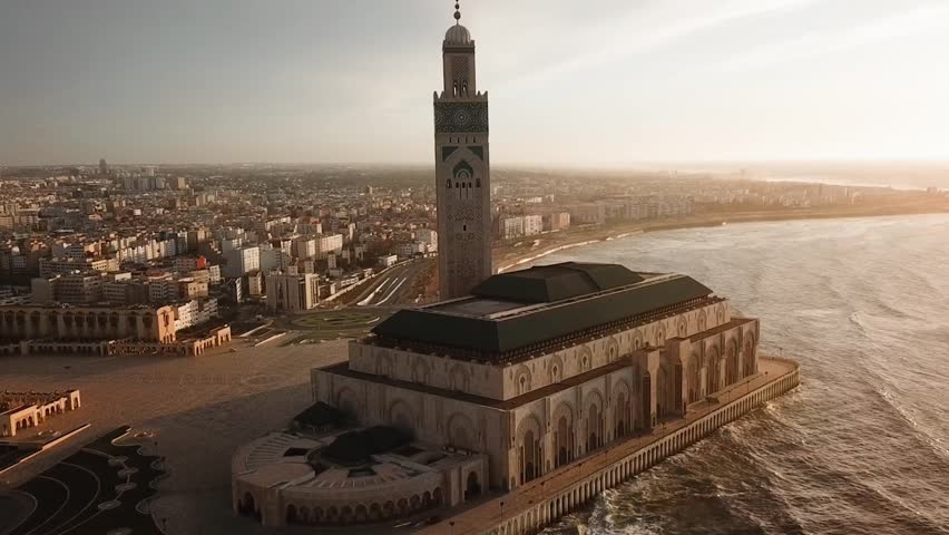 Breathtaking aerial shot of the Hassan II Mosque during a warm sunset. Features the glowing architectural details, the iconic minaret, and the Atlantic waves at the Casablanca waterfront.