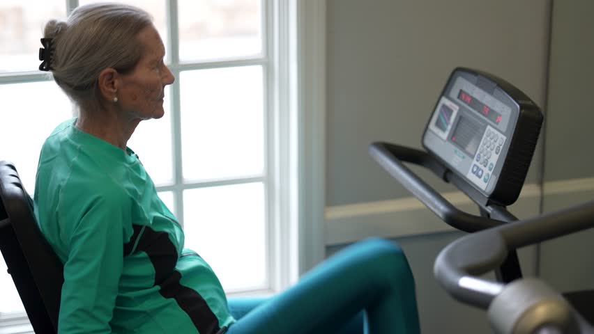 Elderly woman works out on a stationary bike in a gym setting while focusing on her fitness goals in the morning.