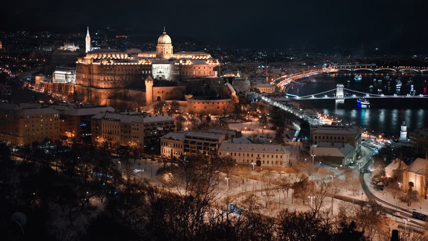 Winter night aerial view of Buda Castle and Chain Bridge illuminated above the Danube River in Budapest, Hungary. Snow covered rooftops and glowing city lights.