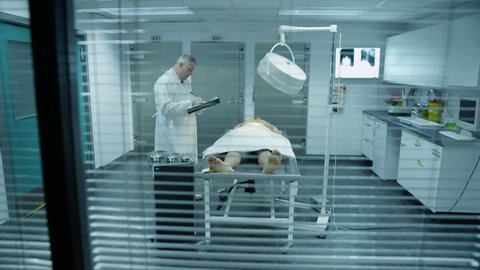 The lifeless naked corpse of a young mixed race male is laid out on the autopsy table, ready for the medical examiner to begin his work. He checks the toe tag and starts to make notes. In slow motion.