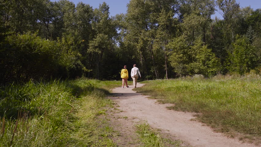 Mom and son walk slowly along path in natural park on sunny day, very wide shot. Family enjoys walk and communication outdoors, comfortable interaction between parent and teenager.