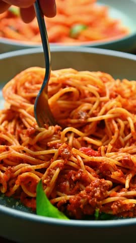 Fork lifting spaghetti pasta noodles with tomato sauce garnish. Macro view of pasta lifted by metal fork over plate with blurred background