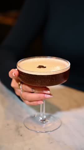 Woman holding espresso martini cocktail with coffee beans garnish. Close up of a person holding a stemmed glass filled with espresso martini topped with foam and coffee beans.