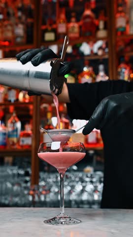 Bartender pouring pink cocktail through strainer into coupe glass. Professional bartender in black gloves pouring drink from shaker through sieve into glass on marble bar counter.