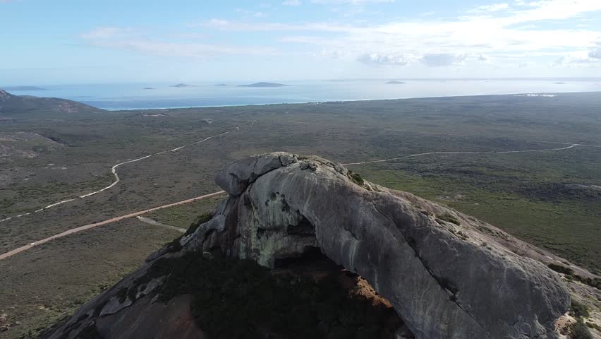 Cinematic drone shot starting almost directly above Frenchman Peak in Cape Le Grand National Park, Western Australia, then flying backward to reveal the full granite dome and surrounding wilderness landscape. Epic aerial perspective showcasing the scale of the isolated mountain against the vast Australian terrain and distant coastline.