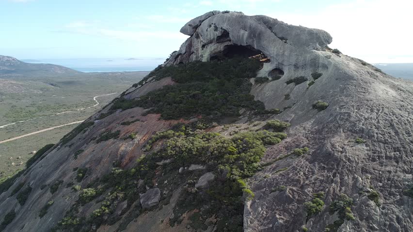 Cinematic drone shot ascending vertically along the granite slopes of Frenchman Peak in Cape Le Grand National Park, Western Australia. As the drone rises, it reveals breathtaking turquoise beaches and expansive coastline stretching across the horizon behind the mountain. Dramatic Australian landscape combining rugged granite formation with pristine ocean scenery.