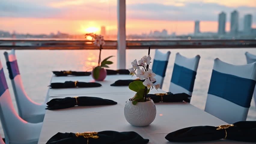 Set dinner table on cruise ship with sunset over water. Restaurant table setting with white orchids and lobster napkin rings on boat with sunset over the horizon.