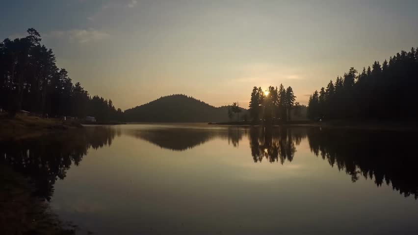 The golden sun peeks through silhouetted pine trees, casting a warm glow over a tranquil lake and its mirrored landscape