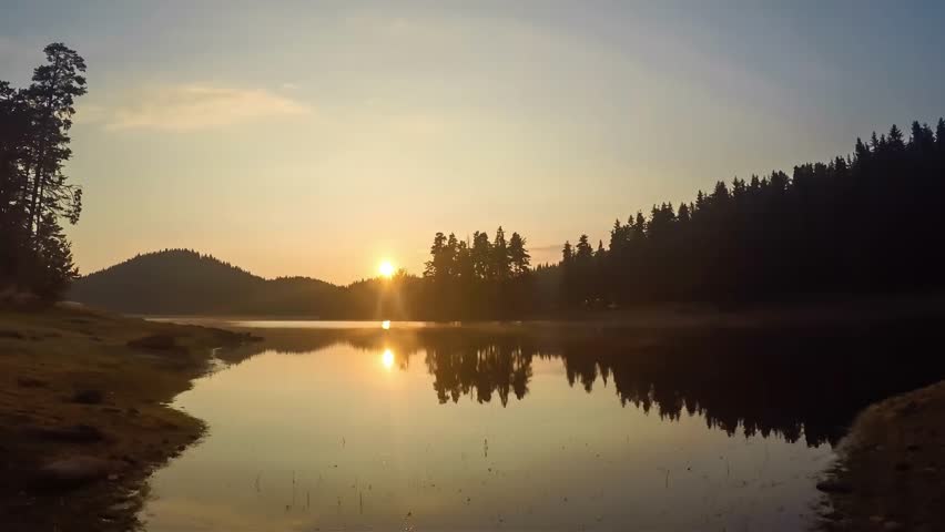 Golden sunbeams break through the morning mist over a tranquil lake, illuminating the surrounding evergreen forest and its perfect reflection