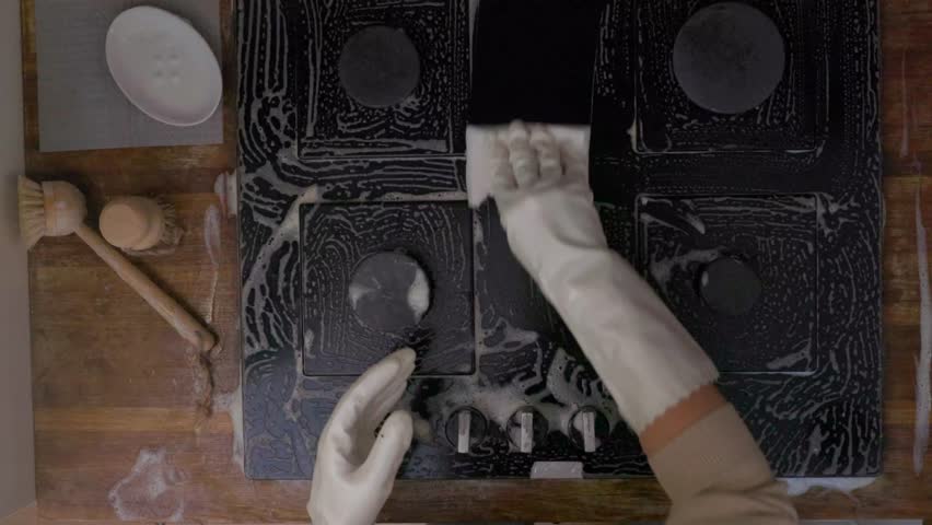 Overhead shot of female hands washing black glass cooktop with soap foam. Domestic cleaning routine, modern kitchen interior