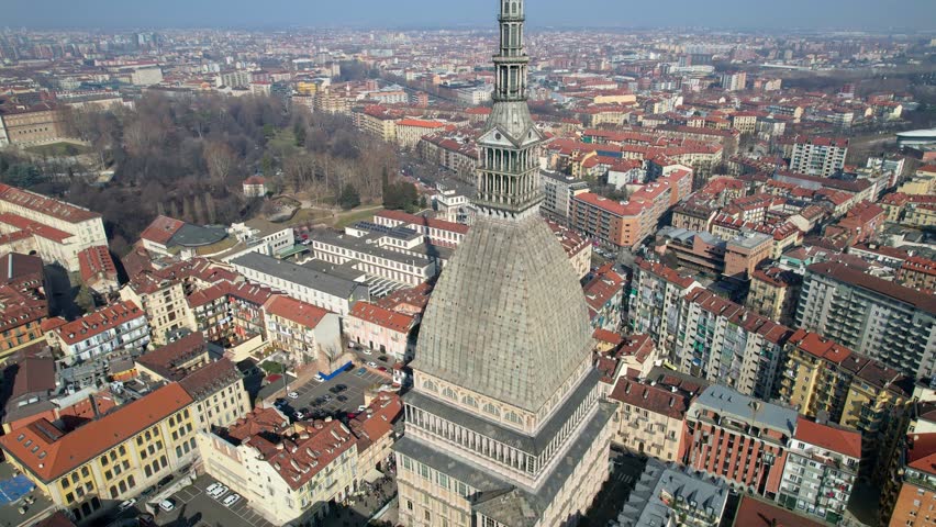 High angle drone shot shows the textured stone dome and spire of Mole Antonelliana overlooking the historic city center of Turin, Italy, urban architecture, aerial city view, historic monument.