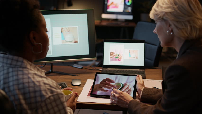 Two biracial colleagues, including photographer and designer, sitting at desk and examining images on tablet and computer