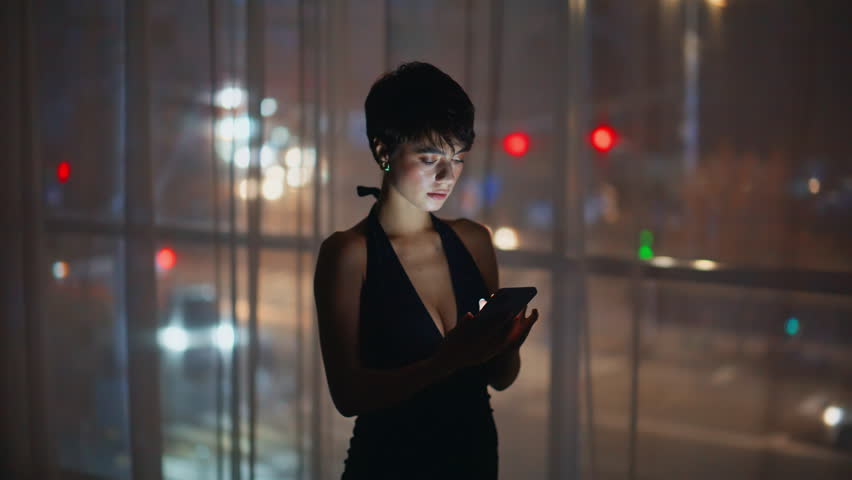 Late brunette texting cellphone standing hotel window closeup. Beautiful sad woman messaging smartphone at night apartment. Depressed lady reading sms notification using mobile phone alone indoors 