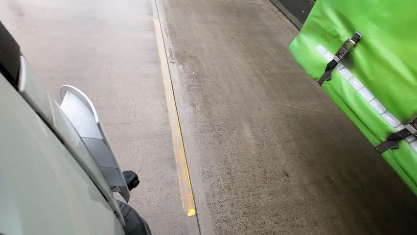 A high-angle side view from a truck cab as it reverses or maneuvers alongside a green curtain-side trailer, showing the triple-axle configuration and yellow floor safety arrows on a concrete loading bay