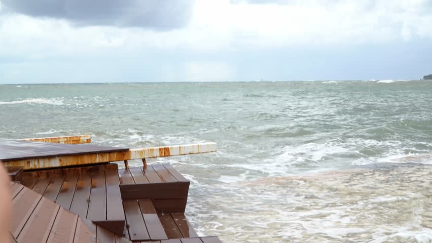 A blonde girl in a black jacket recording the waves of the ocean with her black smartphone.