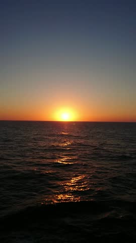 A scenic wide shot of a bright orange sun setting over the calm dark blue water of the Aegean Sea. The camera pans to show the white foam wake of a cruise ship or ferry. Beautiful evening sky at dusk in Greece.