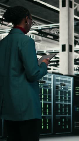 Vertical video Female programmer walking in data center using tablet to write code, compiling software patches. African american woman in server hub using device application to run programming script