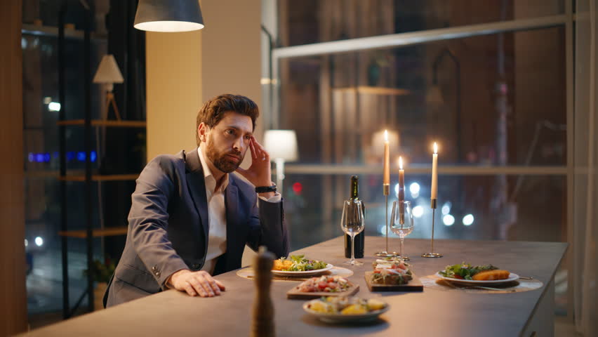 Bored man waiting woman looking wrist watch at hotel table closeup. Sad gentleman sitting alone. Frustrated single guy feeling lonely checking time at cozy apartment. Unhappy brunette guy failed date