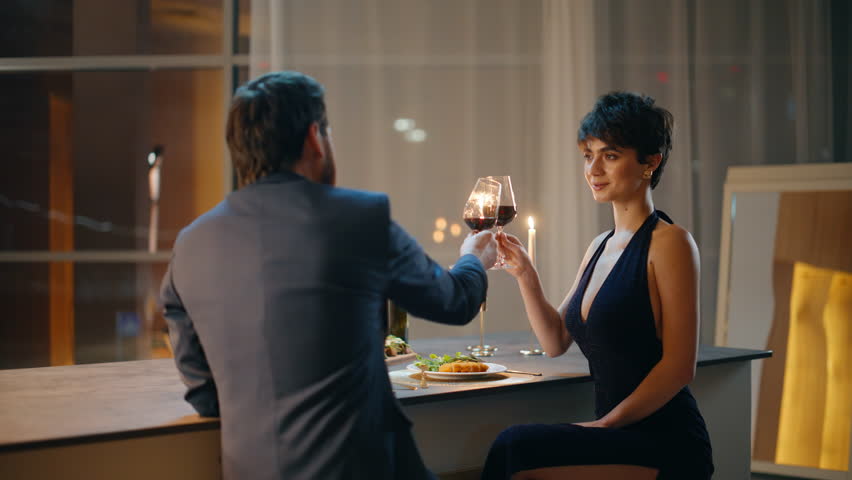 Smiling lovers clinking wine glasses spending evening date sitting bar counter. Loving husband enjoying moment with wife celebrating anniversary luxury modern flat. Happy couple enjoying communication