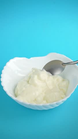 Thick creamy greek yogurt stirred with spoon in white ceramic bowl on blue background.	