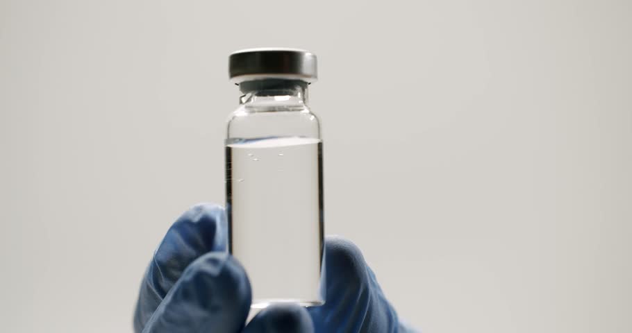 Medical professional in blue gloves holding and shaking a glass vial with a transparent liquid medication, representing a vaccine for injection, pharmaceutical research, or a new drug