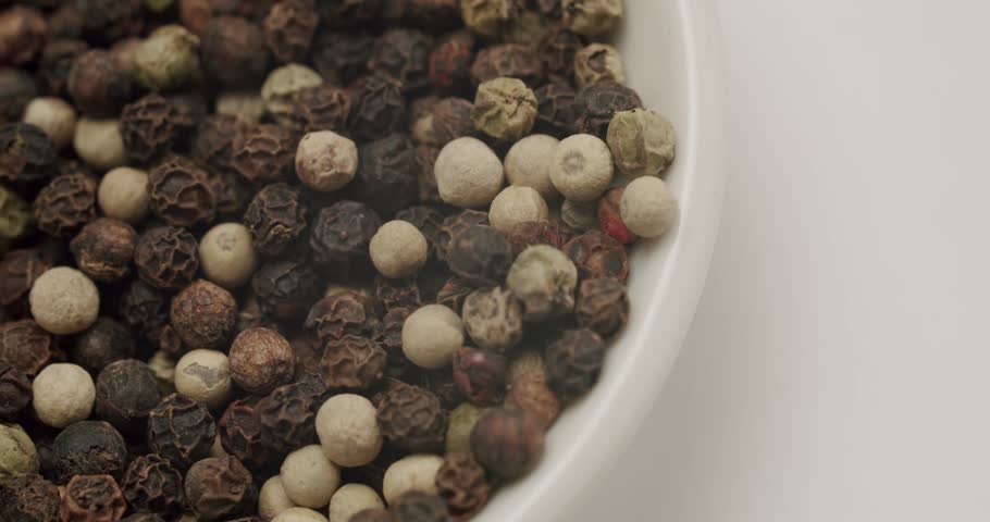 Macro shot of a bowl filled with a colorful mix of black, white, green, and red peppercorns rotating slowly. The aromatic and spicy condiment is isolated on a clean white background