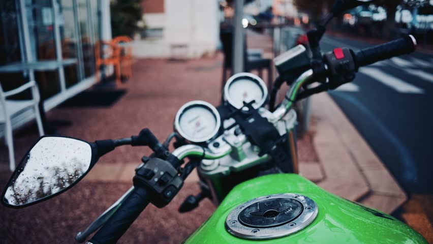 A close up view of the green fuel tank and handlebars of a parked motorcycle with drops of water on the mirror