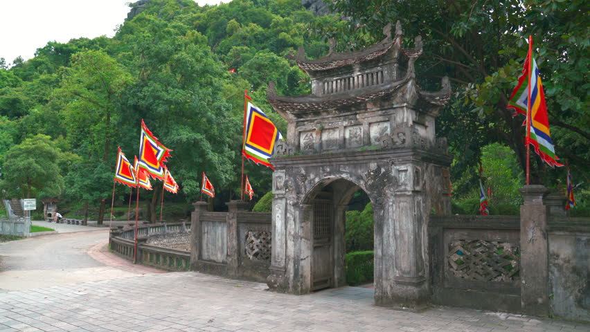 A historic stone entrance gate adorned with vibrant traditional Vietnamese festival flags, leading to the ancient capital complex of Hoa Lu in Ninh Binh.