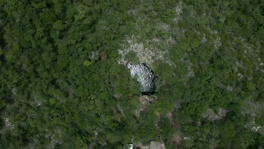 High-altitude top-down aerial ascent revealing an isolated cenote in the middle of the Yucatán jungle. Sky reflections shimmer on the water surface near Mérida, Mexico.