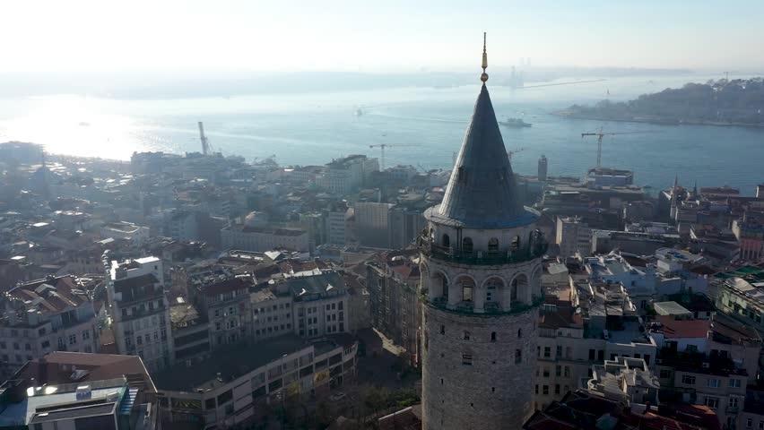 Drone footage showcasing the historic Galata Tower in Istanbul, Turkey. The medieval stone tower with its conical roof rises above a densely packed urban landscape of historical and modern buildings. The Golden Horn waterway and connecting bridges are visible in the background, highlighting the city’s architectural diversity and cultural heritage. Perfect for themes of history, travel, architecture, and iconic landmarks.