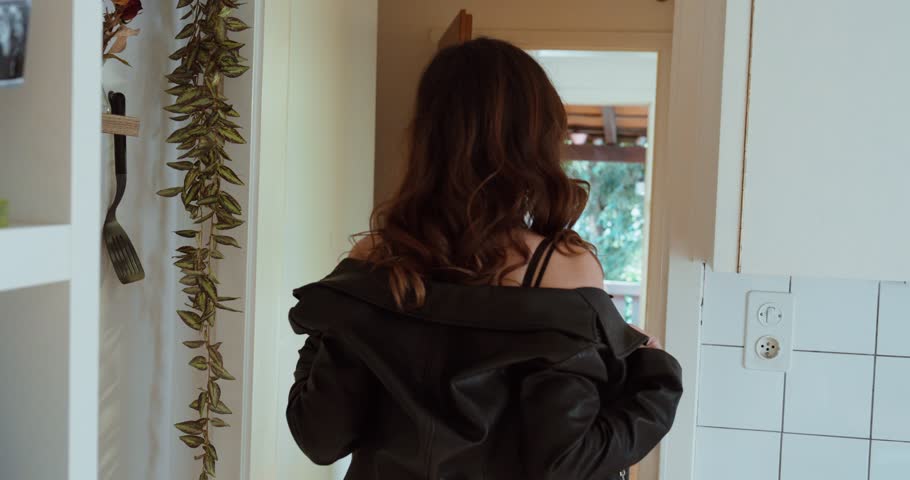 Indoor back view of a woman putting on a leather jacket and walking out the door. Concept of independence, decision, lifestyle movement and everyday departure scene.