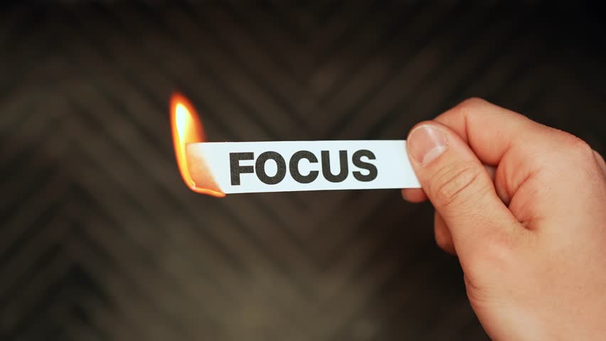 Close-up hand holds a paper label reading “FOCUS” as it catches fire, symbolizing burnout, distraction, and lost productivity on a dark background.