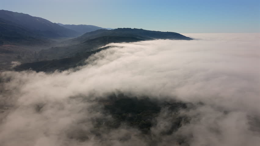 Aerial perspective of Big Sur California mountains rising above thick coastal fog with dramatic ridgelines and green valleys under clear blue sky along Pacific coast.