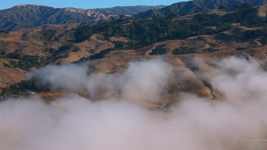 Golden mountain peaks of Big Sur California tower above sea of fog revealing sweeping valleys and rugged Pacific landscape beneath bright clear blue sky.