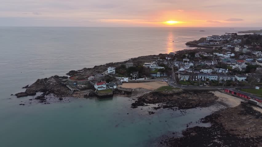 DUBLIN, IRELAND – FEBRUARY 21, 2026: Slow aerial drone push-in toward Sandycove Point at sunrise overlooking Dublin Bay. The footage features the historic James Joyce Tower and Museum, the Forty Foot bathing place, surrounding coastal residences, and Dalkey Island in the background.