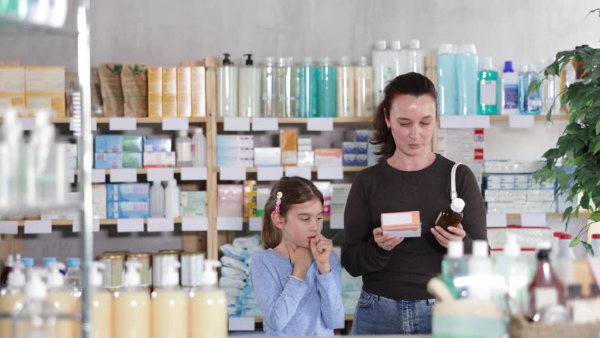 Child stands in the pharmacy with a sore throat and cough, and the mother chooses medicines and syrup for a cold. Daughter and mother came to the pharmacy in search of medicines for colds and flu
